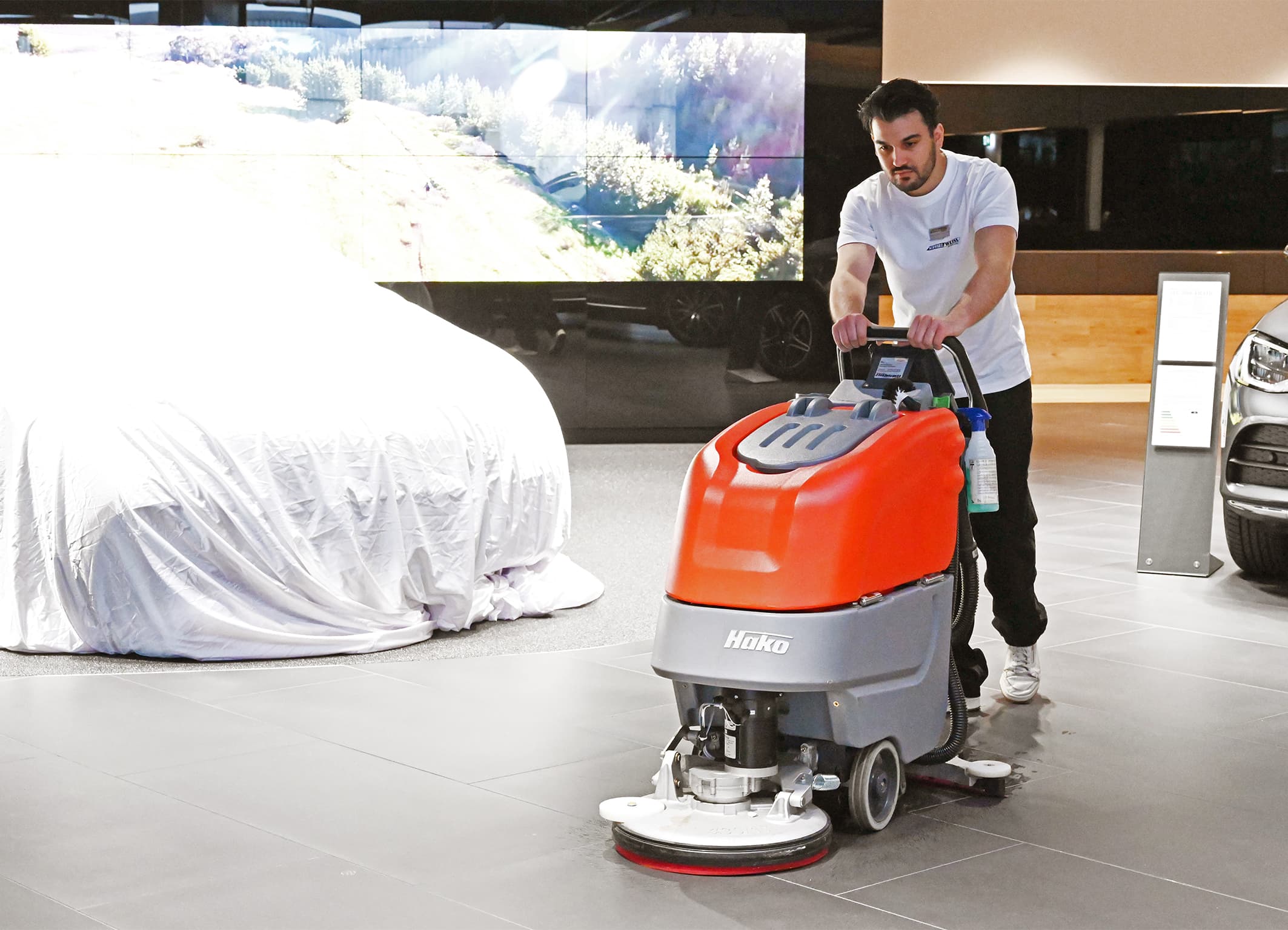 Ein Mitarbeiter reinigt mit einer orangefarbenen Scheuersaugmaschine den Fliesenboden in einem modernen Autohaus.
